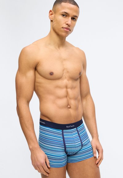 Homme musclé debout, portant un boxer bleu à rayures multiples avec une ceinture de taille marine, regardant légèrement de côté sur un fond clair uni.