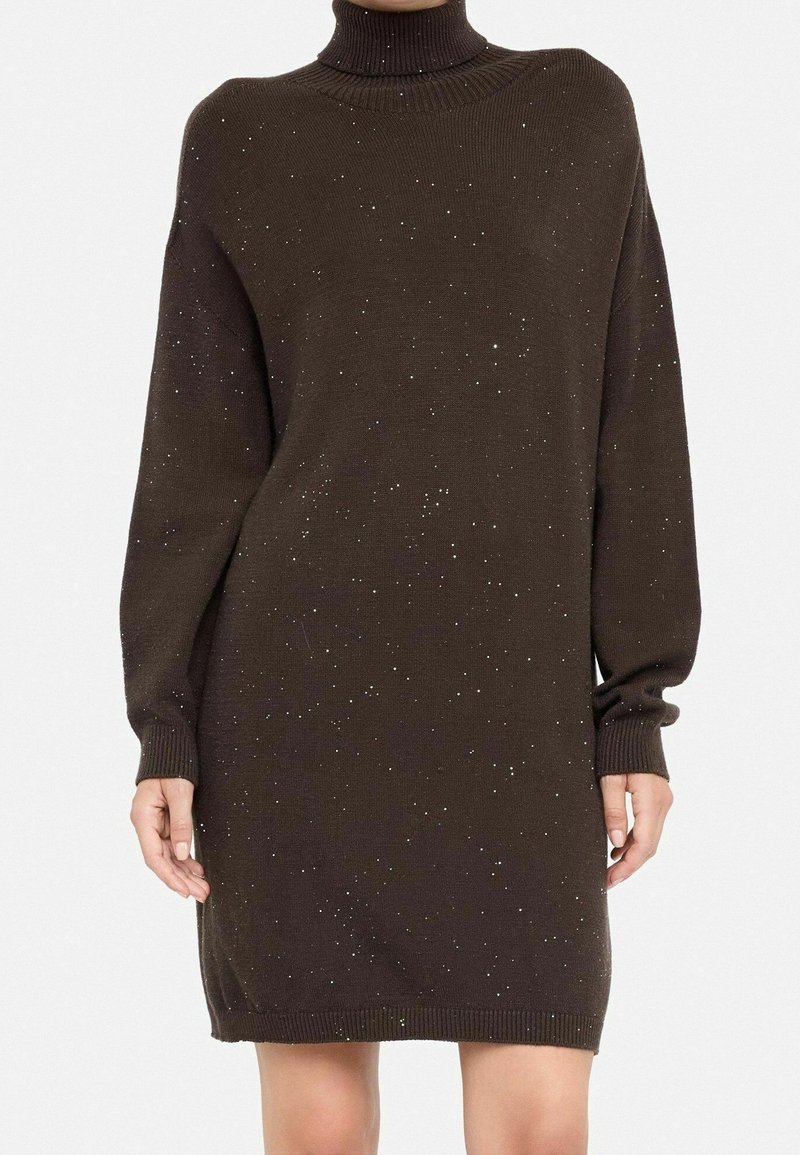 Robe-pull à col roulé marron foncé à manches longues avec un motif moucheté portée par une personne aux mains et jambes découvertes.