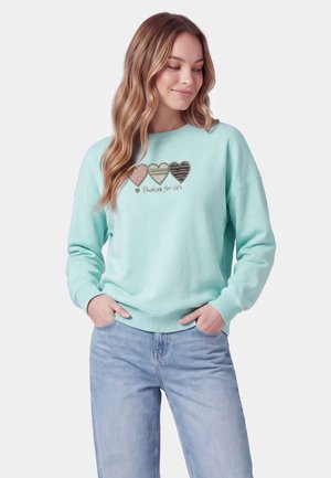 Kvinde iført en lyseblå sweatshirt med tre hjerter og teksten "Pookies for Life", kombineret med lyseblå jeans, hænderne i lommerne.