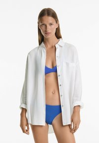 Bela srajca s kratkimi rokavi in sproščenim krojem, zvitih rokavih in žepom; v kompletu z modrim teksturiranim bikini zgornjim delom in usklajenimi hlačkami.