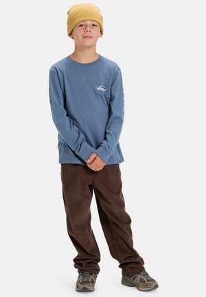 Langarmige blauwe katoenen shirt met grafische details, gecombineerd met een bruine corduroy broek en een gele gebreide beanie. Getextureerde schoenen maken de outfit compleet.