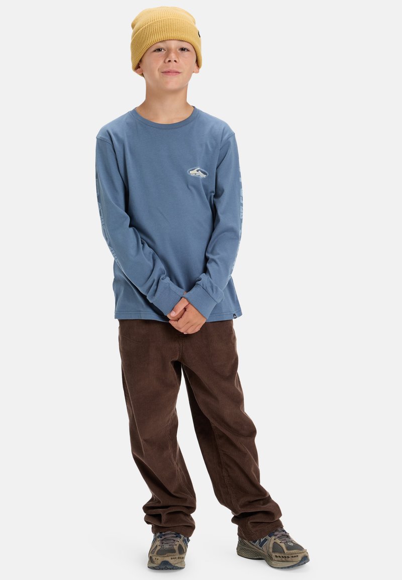 Långärmad blå bomullsskjorta med grafisk detalj, matchad med bruna kordfläskbyxor och en gul stickad mössa. Texturerade skor fulländar outfiten.