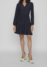 Mörkblå klänning med långa ärmar, V-ringning, diamantmönster, lager på lager-skirt och broderi med hål. Bärs med krämfärgade klackskor.