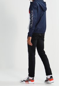 Marinblå vindjacka med huva, röda och vita detaljer samt logotyp på ärmen. Matchad med svarta jeans och flerfärgade sneakers.