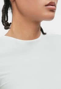 Λευκό βαμβακερό μπλουζάκι με στρογγυλή λαιμόκοψη, λεία υφή, εφαρμοστή σχεδίαση, με ορατά ραφάκια γύρω από τη λαιμόκοψη, που παρουσιάζεται εν μέρει δίπλα σε δέρμα.