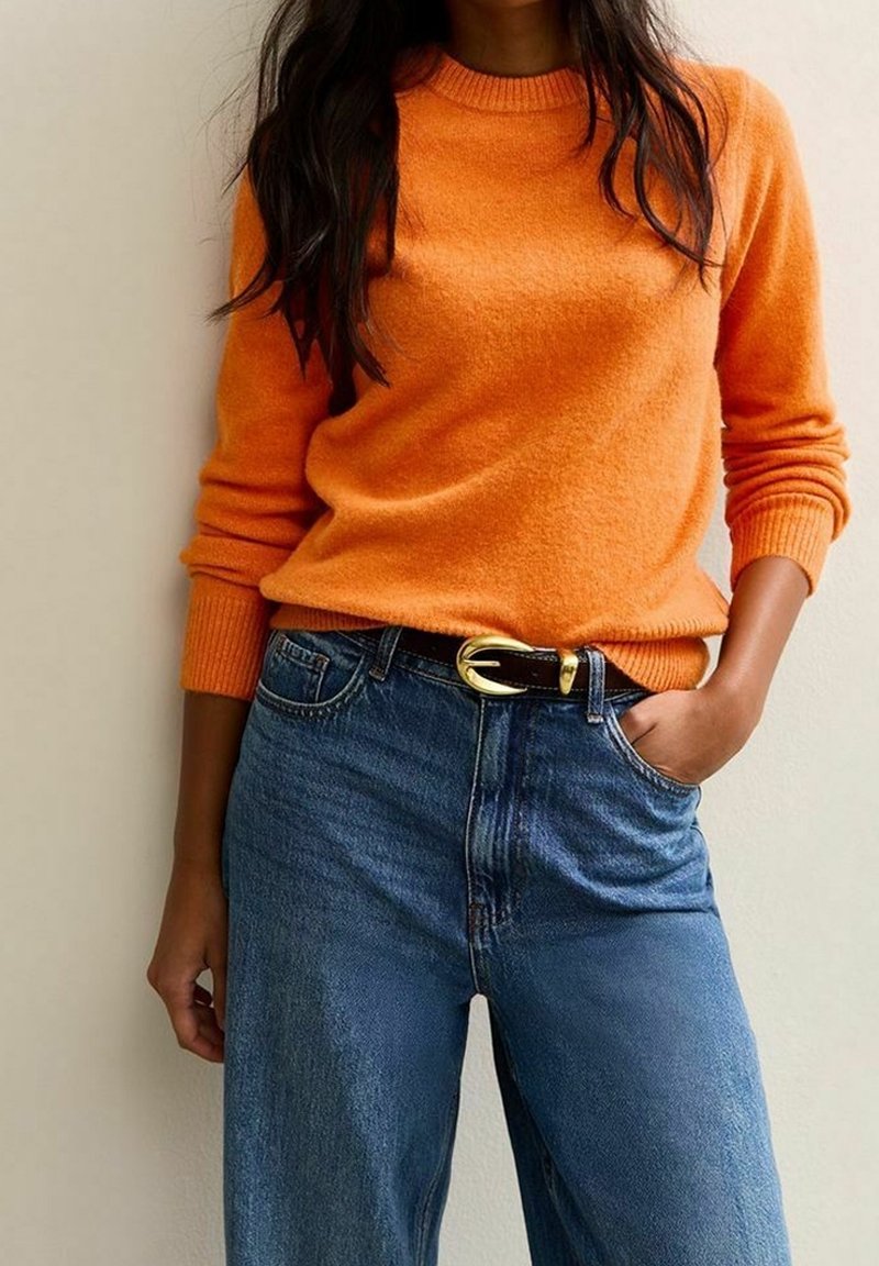 Personne portant un pull orange vif rentré dans un jean bleu taille haute avec une ceinture noire à boucle dorée.