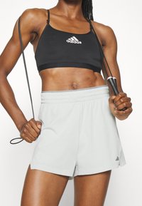 Top corto sportivo nero con spalline sottili e logo Adidas bianco, abbinato a pantaloni corti grigi chiari con vita elasticizzata e una texture sottile.