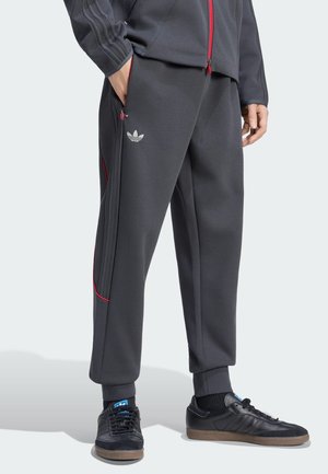 Persoon draagt donkergrijze Adidas trainingsbroek met rode bies en zwarte Adidas sneakers, staat met één hand in de zak.