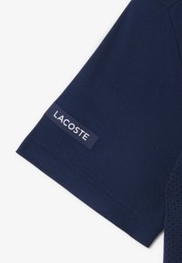 T-shirt bleu marine à manches courtes avec un patch en tissu texturé portant le logo blanc "LACOSTE" près de l'ourlet.