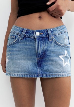 Denim-Minirock mit silbernen Nieten und weißen Sternmotiven, kombiniert mit einem schwarzen Top, das den Bauch zeigt, und einer Hand, die den Bund anpasst.
