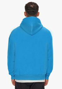 Blaues Kapuzensweatshirt mit weichem Material, lockerer Passform und gerippten Bündchen; von hinten gezeigt, mit einem darüber getragenen weißen Hemd.