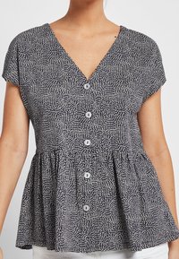 Kvinde iført en sort-hvid mønstret kortærmet bluse med V-udskæring og knapper nedad.