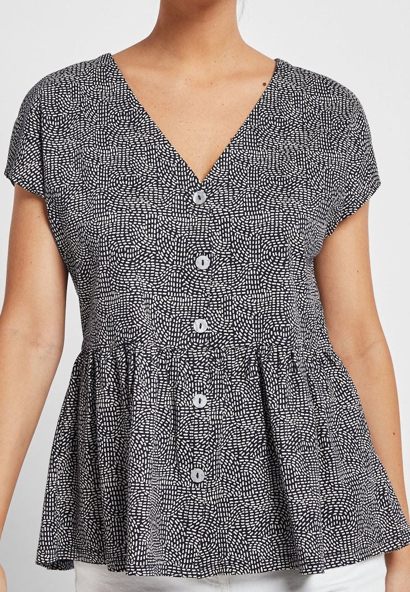Kvinde iført en sort-hvid mønstret kortærmet bluse med V-udskæring og knapper nedad.