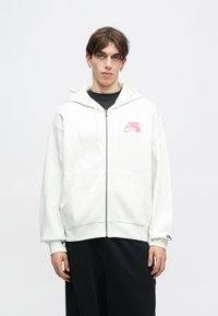 Sweatshirt branco com fecho e bolsos frontais, apresentando o logo rosa da Nike SB no lado esquerdo do peito. Tecido suave e capuz com cordão.