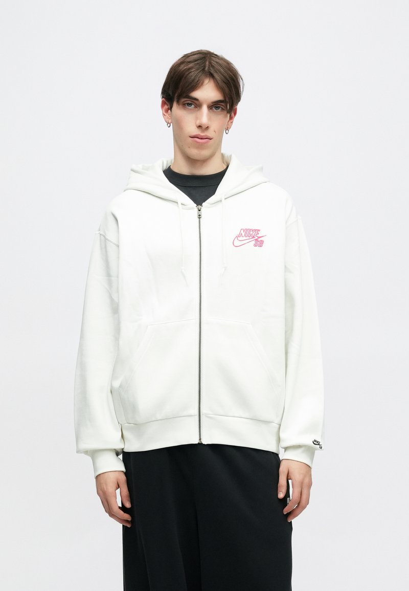Sweatshirt branco com fecho e bolsos frontais, apresentando o logo rosa da Nike SB no lado esquerdo do peito. Tecido suave e capuz com cordão.