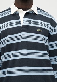 Μακρυμάνικη ριγέ ρουγκμπί μπλούζα σε σκούρο και ανοιχτό μπλε με λευκό γιακά και πράσινο λογότυπο κροκόδειλου στο στήθος.