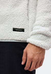 Gros plan d'une main à côté d'une veste polaire blanche texturée avec une étiquette noire "Peak Performance" et un pantalon noir.