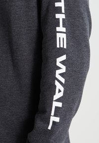 Tummanharmaa pitkähihainen paita, jossa valkoinen "THE WALL" -teksti painettuna pystysuunnassa oikeaan hihaan, valmistettu pehmeästä, teksturoidusta materiaalista.