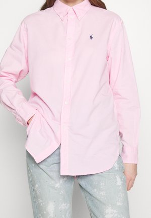 Person trägt ein hellrosa Hemd mit Knöpfen und einem kleinen dunklen Logo auf der Brust sowie hellblaue, mit Farbspritzern versehene Jeans, eine Hand in der Tasche.