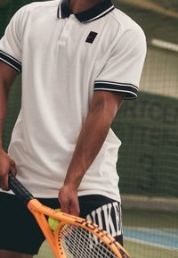 Mies valkoisessa poolopaidassa ja mustissa shortseissa pitää oranssia tennis-mailaa ja palloa ulkoisella tennis kentällä, taustalla verkko.