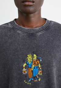 Grijze sweatshirt met kleurrijk geborduurd ontwerp van twee cartoonfiguren die voorwerpen vasthouden. Geribde kraag en textiele stof.