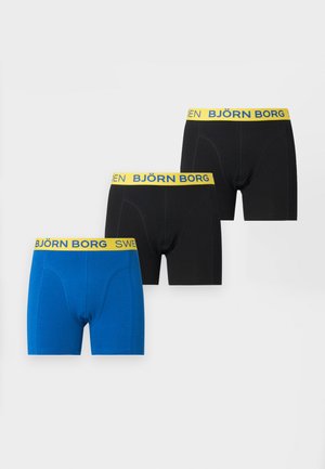 Štíhly svalnatý čierny muž stojaci naboso, oblečený v čiernych boxerkách so žltým pásom, na ktorom je nápis "SWEDEN BJÖRN".