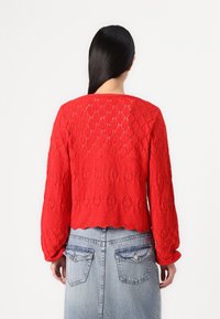 Suéter vermelho tricotado com um padrão texturizado de diamantes, gola redonda e bainha ligeiramente ondulada. Mangas longas e bufantes.