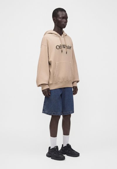Homme portant un sweat à capuche beige Off-White, un short en jean bleu, des chaussettes blanches et des baskets noires, debout devant un fond uni.