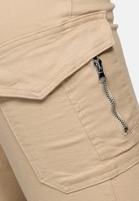 Pantalones cargo beige con un bolsillo lateral con cremallera, tejido texturizado y costuras reforzadas. El diseño incluye una solapa sobre el bolsillo.