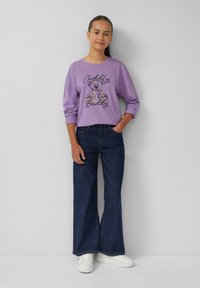 Lila sweatshirt met de opdruk "Cuddly Buddy" met daarop een beer, gecombineerd met donkere wijde jeans en witte sneakers. Gladde textuur, casual stijl.