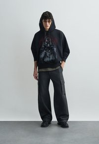 Černá mikina s grafickým potiskem, spárovaná s volnými tmavými cargo kalhotami a černými botami. Model nosí sluneční brýle a stojí proti neutrálnímu pozadí.