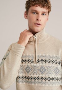 Beżowy sweter z dzianiny z wysokim kołnierzem, z zamkiem błyskawicznym i geometrycznym wzorem w jasnym niebieskim, szarym i czarnym na wysokości klatki piersiowej.