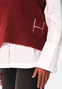 Rød, fluffy strikket vest lagt over en hvit skjorte med knapper. Har broderte, hvite bokstaver "H" på vesten og ermene, med en avslappet passform.