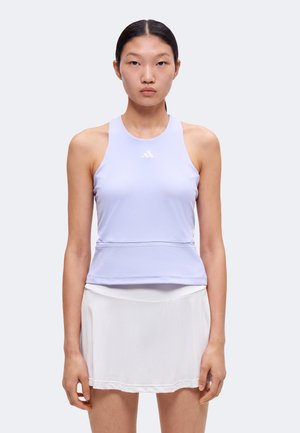 Femme vêtue d'un haut de sport sans manches violet clair et d'une jupe de tennis blanche, debout, les bras détendus, devant un fond clair uni.