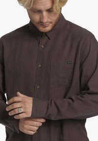 Brown long-sleeve shirt made of textured fabric, featuring a button-down collar, front pocket, and brown buttons for closure.