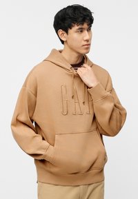 Νεαρός άνδρας που φοράει ανοιχτόχρωμο φούτερ GAP, με το ένα χέρι στην τσέπη και το άλλο να ρυθμίζει το κορδόνι του φούτερ, κοιτάζοντας προς το πλάι.