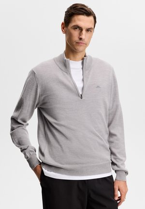 Mężczyzna ubrany w jasnoszary sweter z krótkim zamkiem na przodzie, nałożony na białą koszulę, z czarnymi spodniami, stojący z jedną ręką w kieszeni na jednolitym tle.