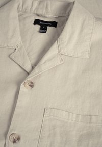 Chemise en lin beige avec col, deux boutons et une poche poitrine gauche. Présente des coutures apparentes et une étiquette de marque portant l'inscription "Matinote".