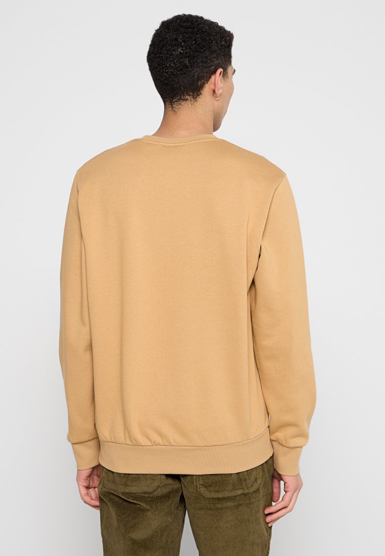 Uomo di spalle che indossa una felpa beige a maniche lunghe e pantaloni in velluto a coste verde oliva su uno sfondo bianco uniforme.