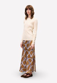 Crème ribbeltrui gecombineerd met een bruine maxi rok met een patroon en blauwe accenten, voorzien van zijsplitten. Zwarte flats maken de outfit compleet.