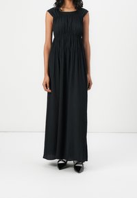 Black maxi dress with vertical pleats, cap sleeves, and a gathered waist. Made of lightweight fabric, paired with pointed black shoes.