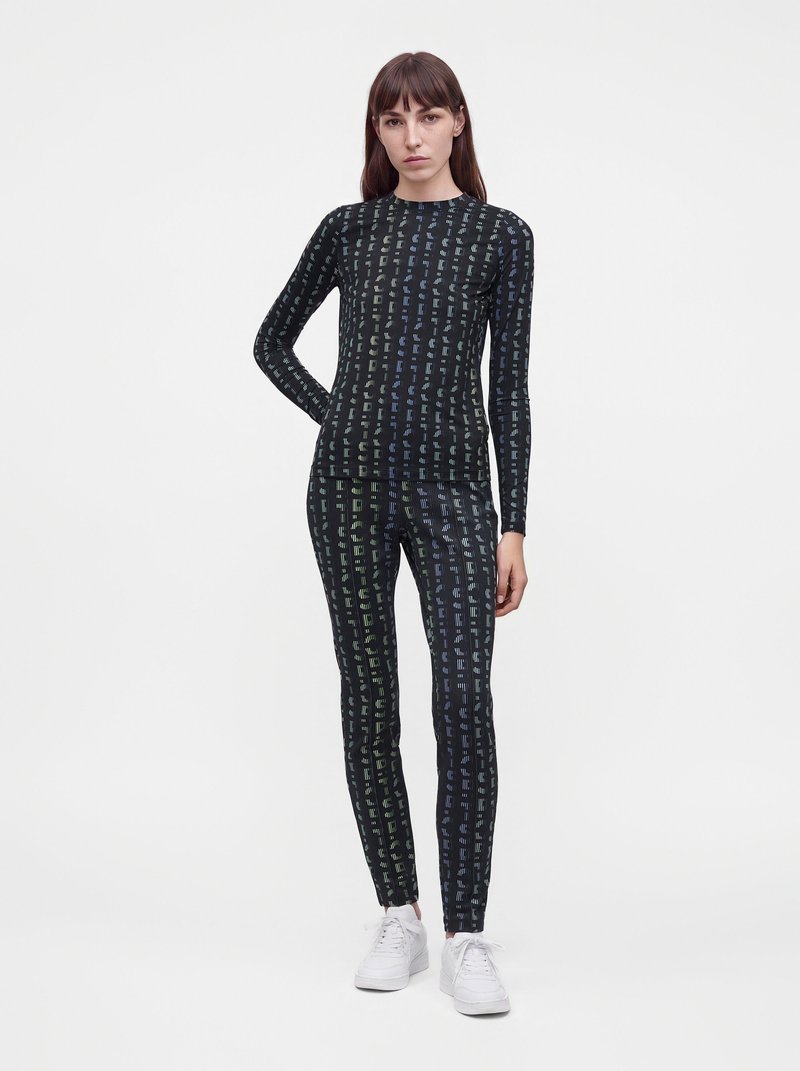 Femme portant un haut noir à manches longues assorti et des leggings avec un motif abstrait vertical, associés à des baskets blanches, debout sur fond blanc.