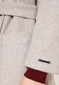 Beige wollen jas met een textuurafwerking, voorzien van een ceintuur in de taille en een rode metallic geribbelde manchet. Zwarte label met witte letters zichtbaar.