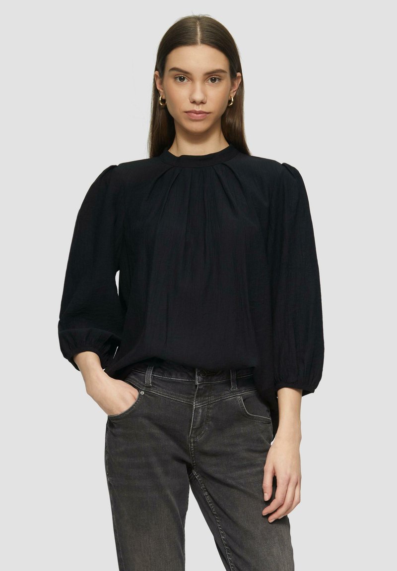 Schwarze Bluse mit dreiviertel langen Puffärmeln, gefaltetem Vorderteil und hohem Ausschnitt. Der Stoff wirkt leicht und leicht strukturiert.