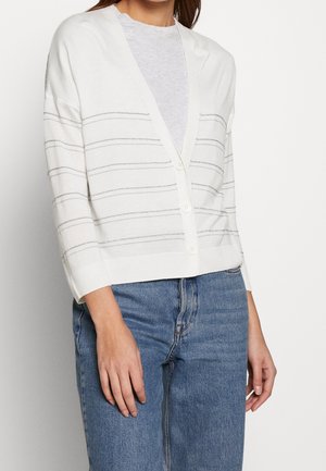 Femme portant un cardigan blanc boutonné avec de fines rayures horizontales sur une chemise gris clair et un jean bleu, debout devant un fond uni.