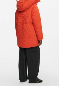Chaqueta acolchada naranja con capucha, con una textura ondulada. Combinada con pantalones cargo negros de corte ancho y zapatillas negras.