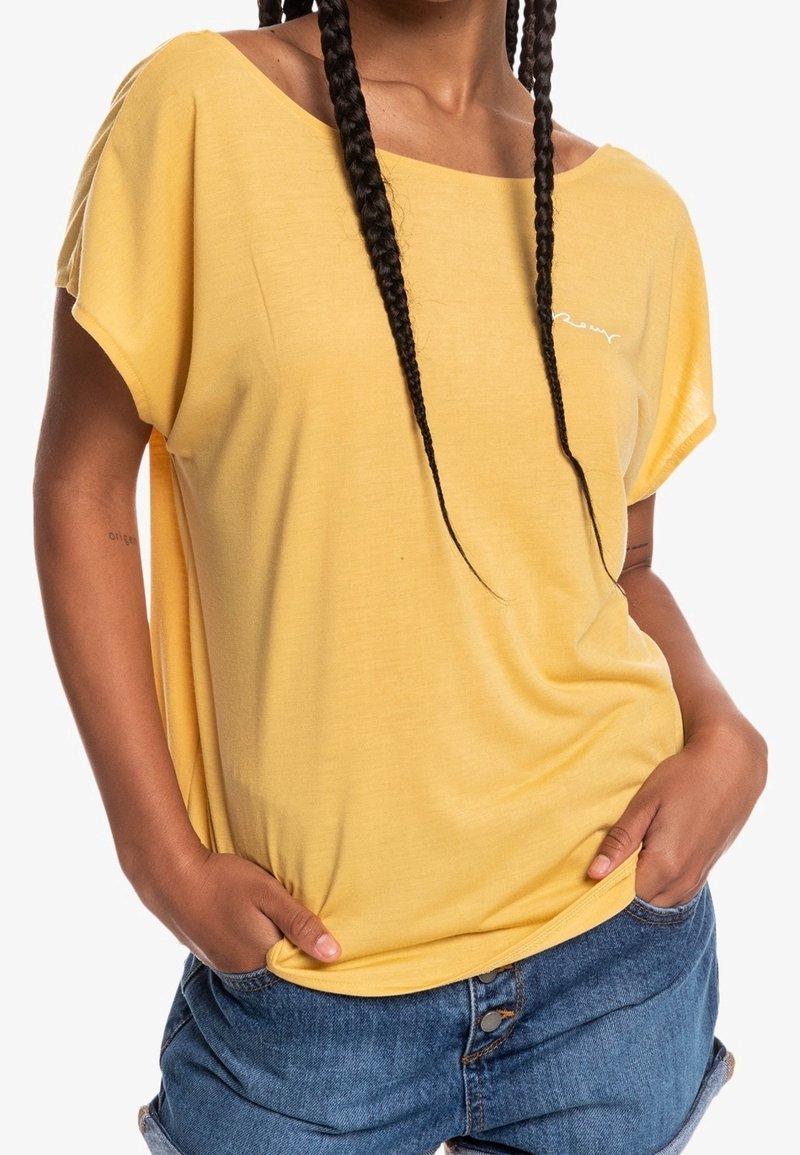 Keltainen, löysä t-paita, jossa on leveä pääntie, lyhyet hihat ja pehmeä rakenne. Edessä pieni valkoinen logo. Yhdistetty farkkushortsien kanssa.