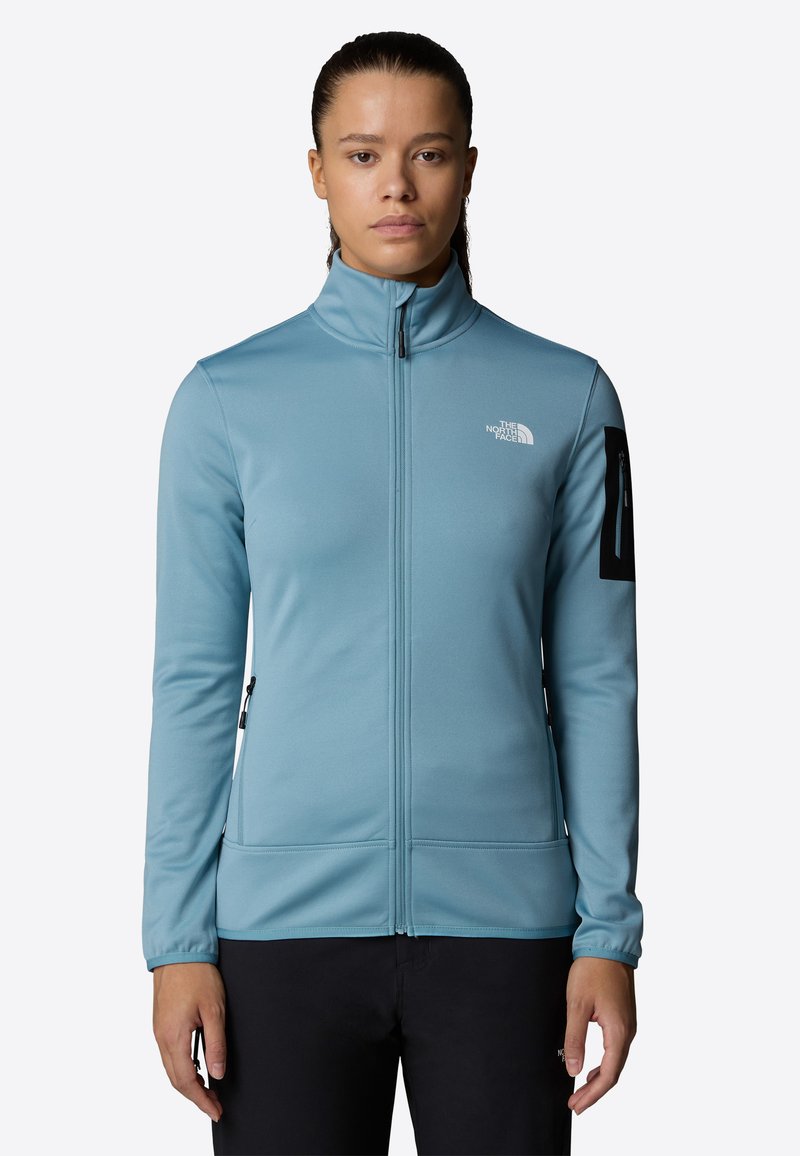 Ljusblå fleecejacka med hög krage, hel dragkedja och svart dragkedjeficka på armen. Slät textur och åtsittande design för värme.