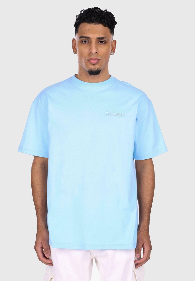 Camiseta de algodón azul cielo con cuello redondo, mangas cortas y un sutil logo bordado en el lado izquierdo del pecho. Corte holgado y textura suave.