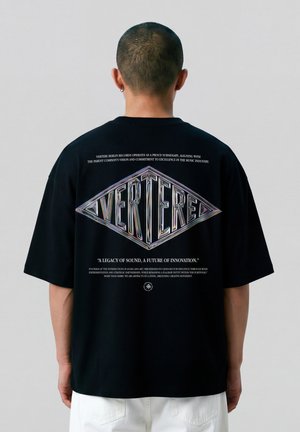 Man draagt een zwart oversized T-shirt met kleurrijk "VERTERER" logo en witte tekst op de achterkant, gecombineerd met witte broek, met de rug naar de camera toe.